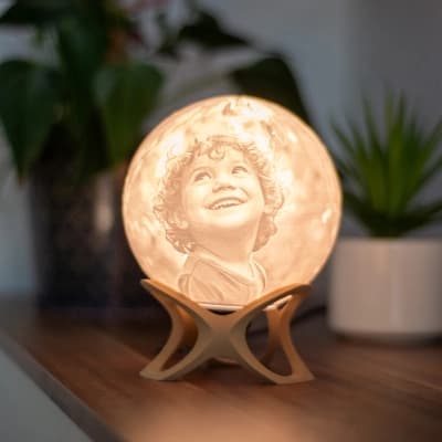 Luna personalisierte 3D-Fotolampe mit einem Kinderporträt, das auf einem Holzsockel leuchtet, auf einem Regal mit Pflanzen platziert.